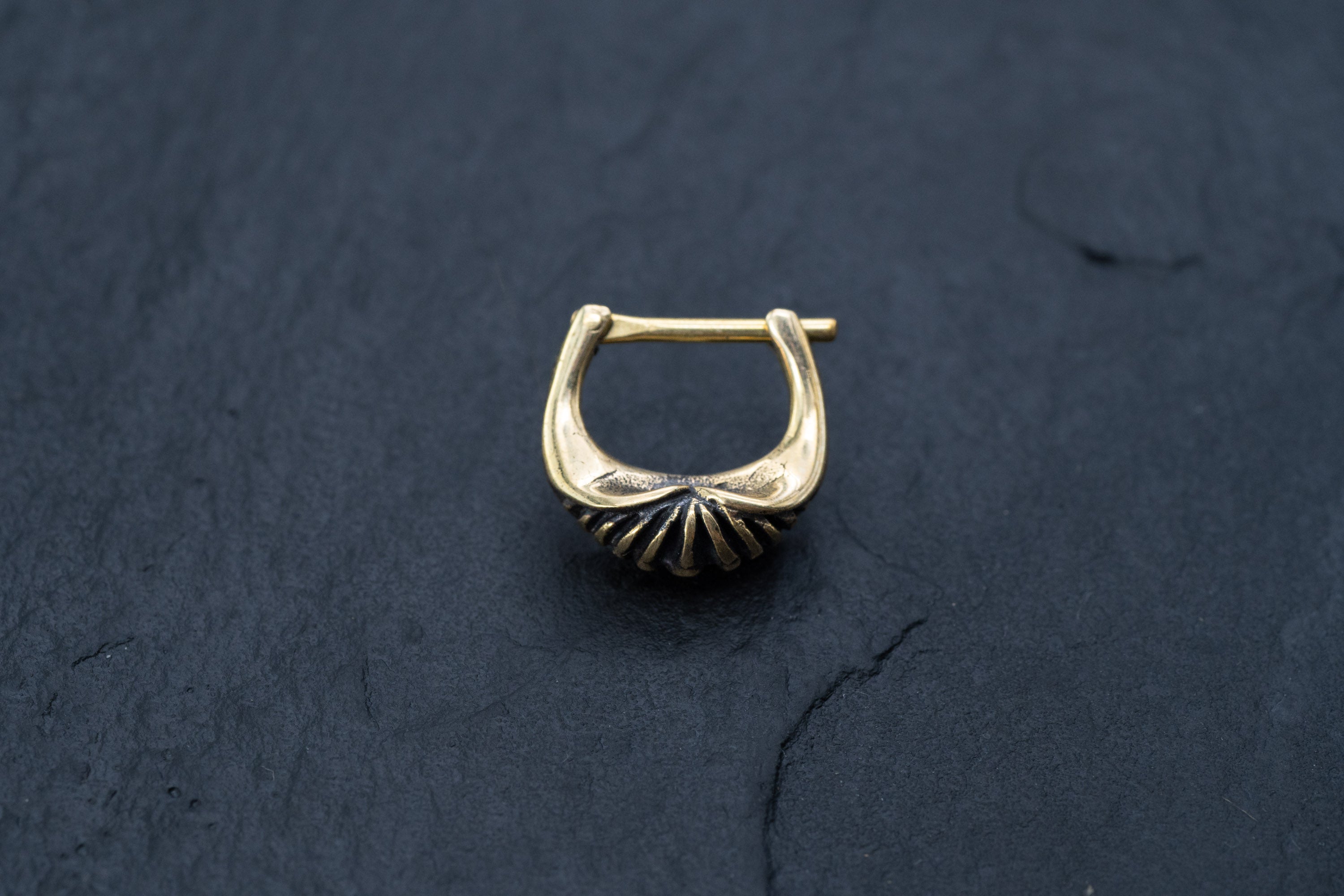 A close-up of the DIARA Biomechanical Clicker Hoop Septum Nose Ring in Gold by Kaya Movement, placed on a dark, textured surface. The hoop earring boasts a unique U-shaped design with a ridged, fan-like accent at its base, reminiscent of a chic unisex nose piercing.